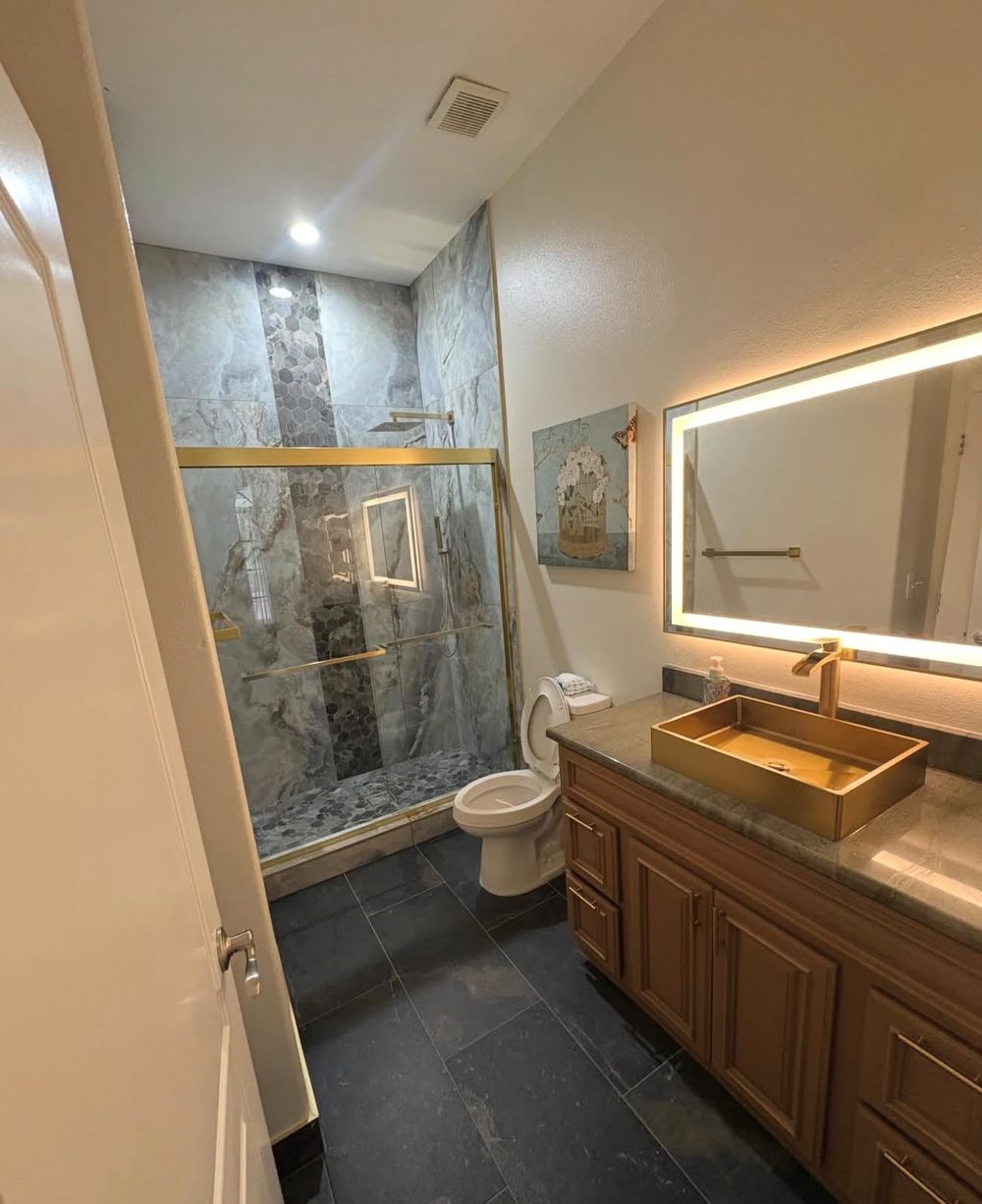 Bathroom with marble-look shower, hexagon accent stripe, brushed gold vessel sink and backlit mirror